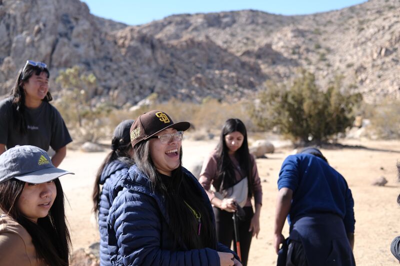 A group of people are gathered in a desert landscape with mountains in the background. The focus is on a woman in a Padres baseball cap who is laughing. Other people are standing around her, some looking in different directions. The overall impression is one of an outdoor excursion or gathering in a natural setting.